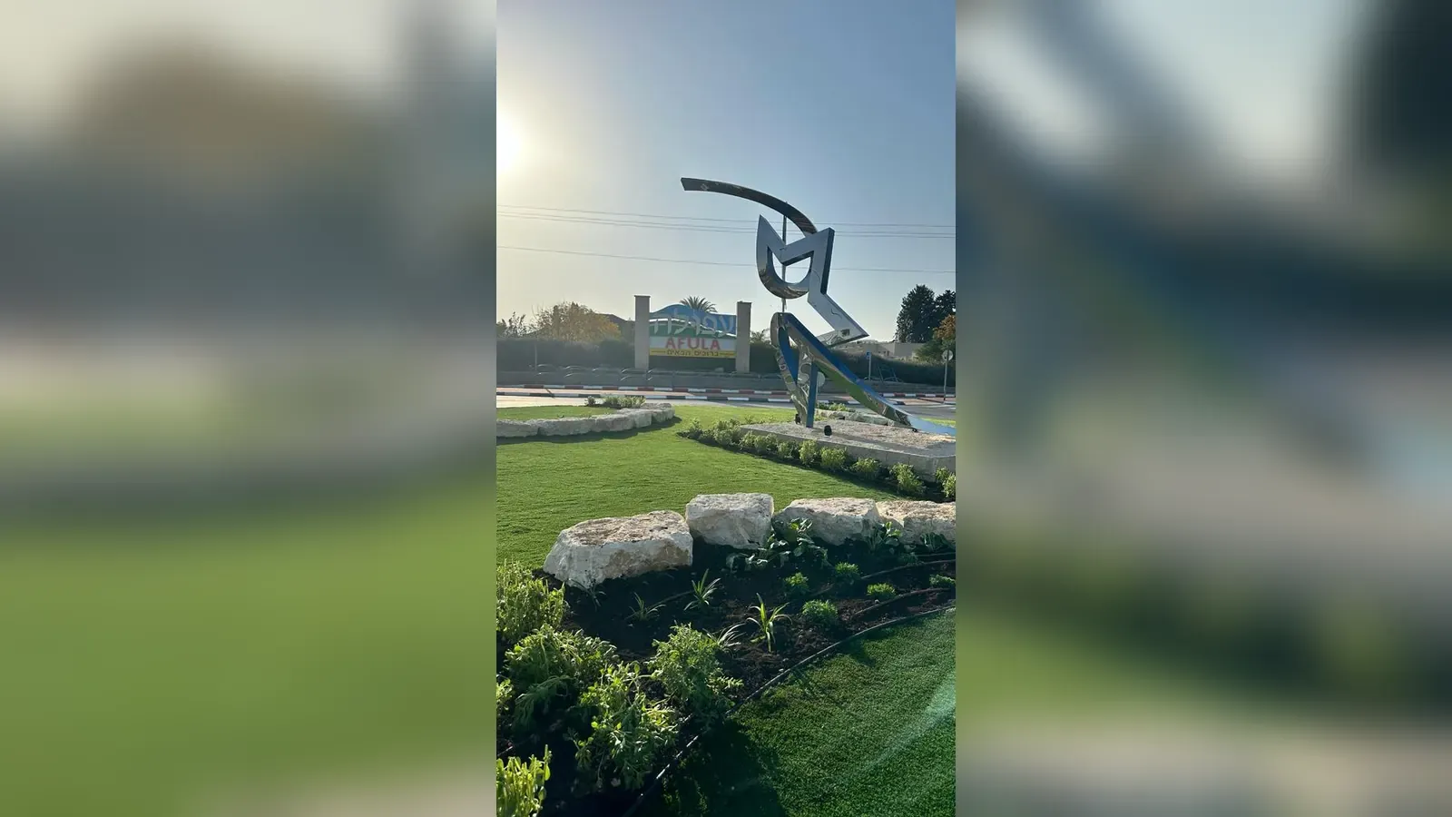 פסל בכניסה לעפולה לצד שילוט העיר, מוקף מסלעה, דשא וצמחייה